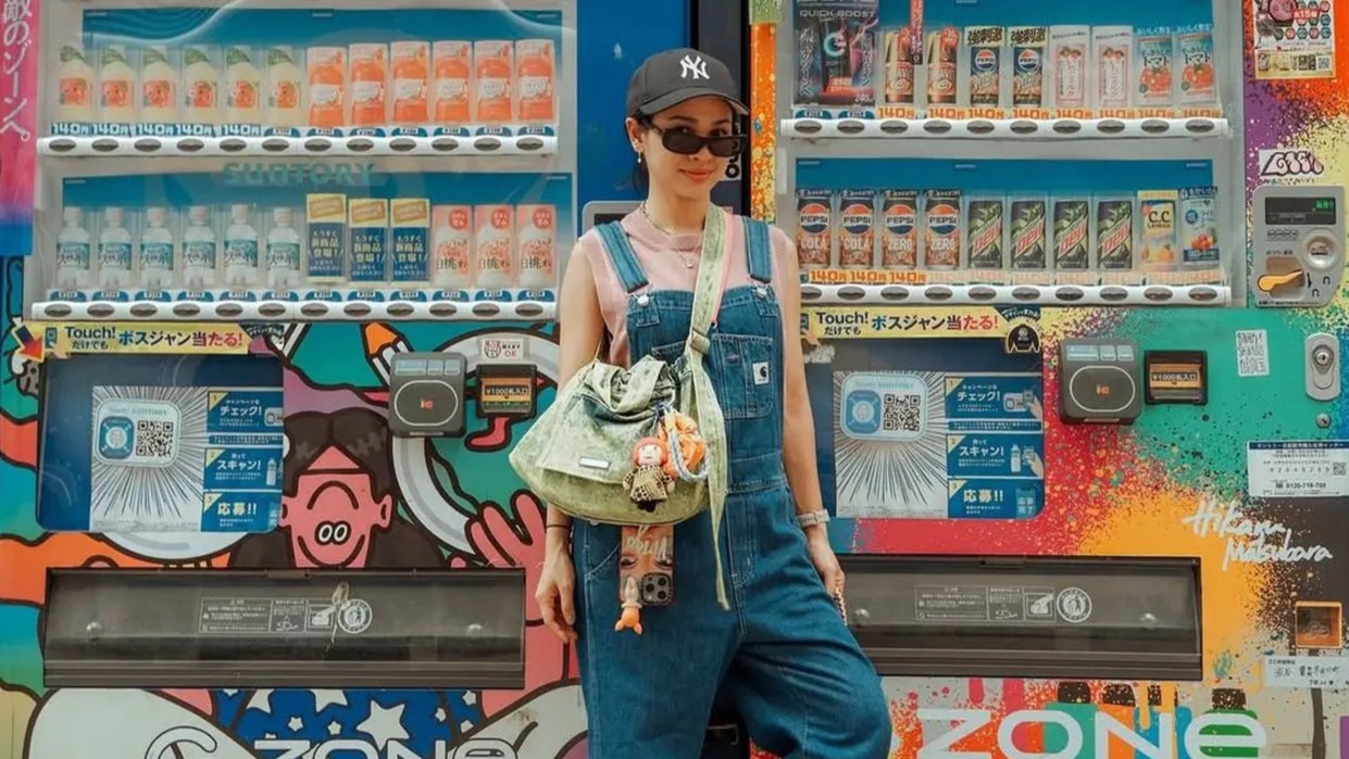 Andien Aisyah tampil kasual chic dengan overall denim, topi baseball, dan tas selempang unik di depan mesin minuman Jepang.