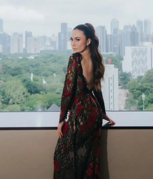Wulan Guritno mengenakan gaun transparan hitam dengan motif mawar merah dan gaya rambut ponytail saat berpose di depan jendela dengan latar kota Jakarta.