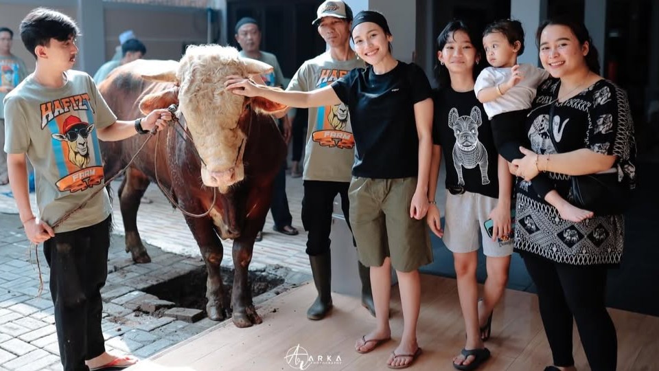 Ayu Ting Ting mengenakan kaos hitam dan celana hijau army saat berpose bersama keluarga dan sapi kurban.