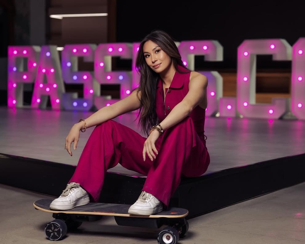 Lucy Guo mengenakan jumpsuit fuchsia duduk di atas skateboard dengan latar dekorasi lampu.