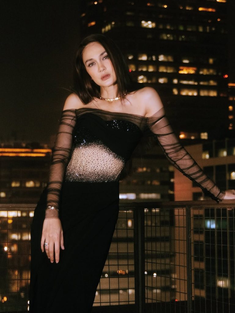 Luna Maya tampil elegan dengan gaun hitam transparan berdiri di balkon menghadap pemandangan kota malam hari