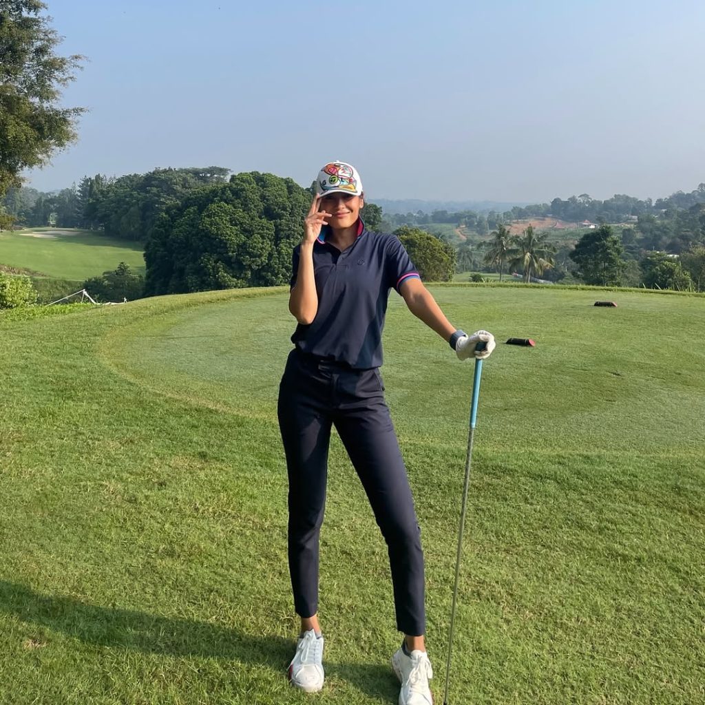 Sigi Wimala mengenakan polo navy dan celana hitam, berdiri di lapangan golf sambil memegang stik.