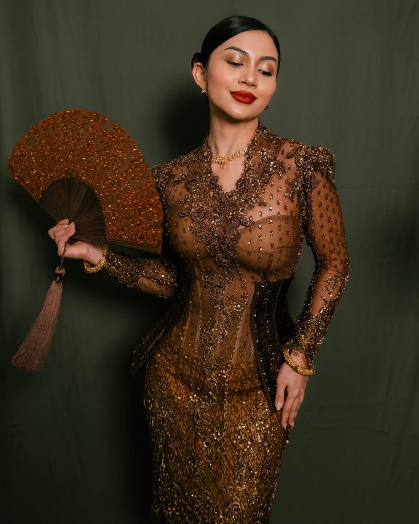 Ariel Tatum mengenakan kebaya cokelat dengan bordiran emas dan waist bustier manik-manik, berdiri anggun di depan latar dekorasi etnik.