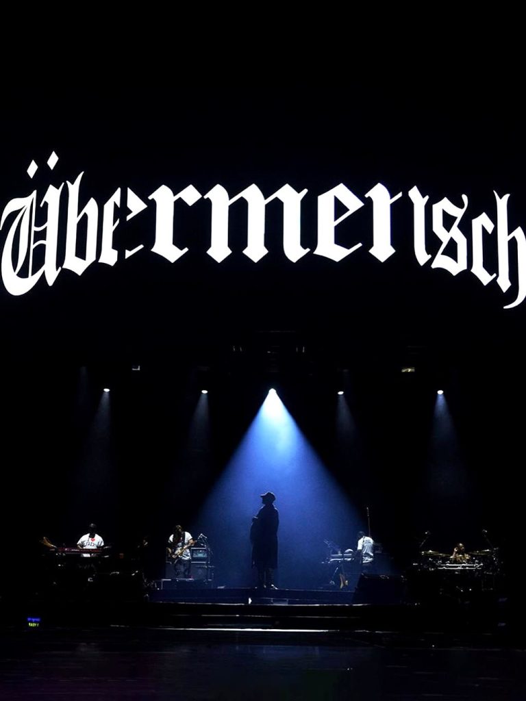 G-Dragon tampil energik di atas panggung konser Übermensch, mengenakan kostum futuristik dan pencahayaan dramatis.