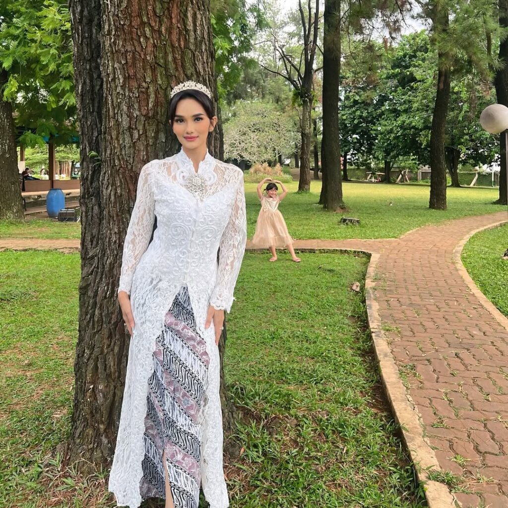 Kebaya putih panjang dengan motif renda yang detail dipadukan dengan kain batik bernuansa hitam-pink membuat penampilannya semakin menawan.