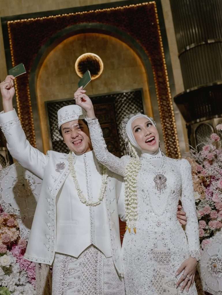Arbani Yasiz dan Raissa Ramadhani mengangkat buku nikah sambil tersenyum lebar di depan dekorasi bunga indah.