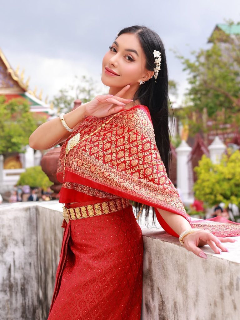Denada tampil cantik di kuil Thailand mengenakan busana tradisional merah keemasan, berpose anggun dengan senyuman.