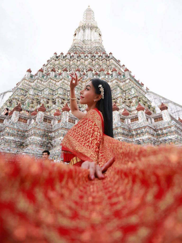 Denada berpose dramatis di depan Wat Arun, membentangkan kain busana tradisional merah emas dengan latar kuil.