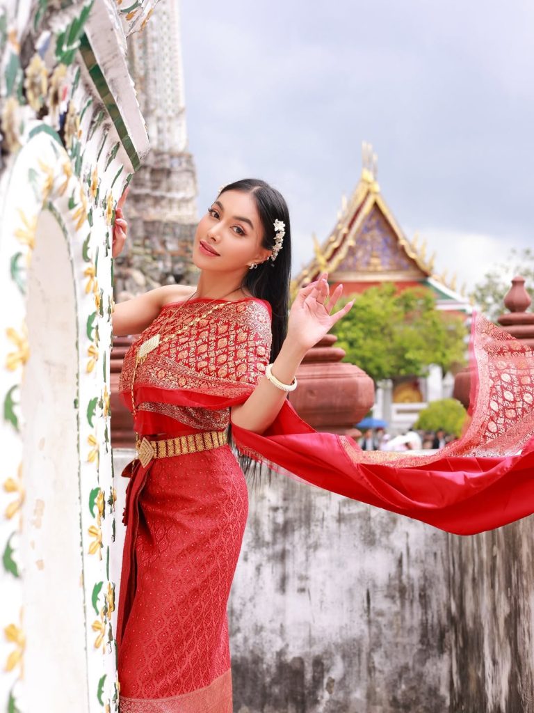 Denada bersandar di pilar kuil Thailand, menatap kamera dengan ekspresi lembut dan busana merah emas menyatu dengan latar.