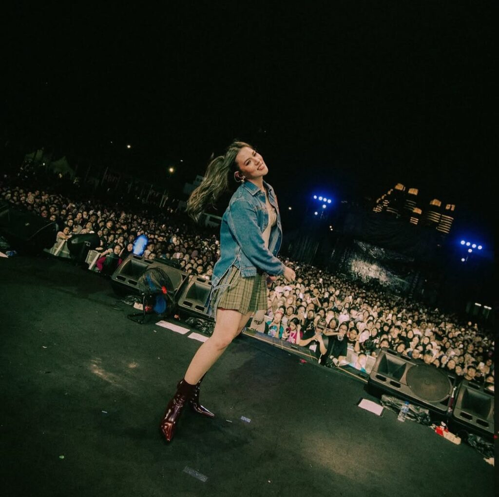 Raisa mengenakan jaket denim dan rok kotak saat tampil di atas panggung dengan senyum lebar di hadapan ribuan penonton)