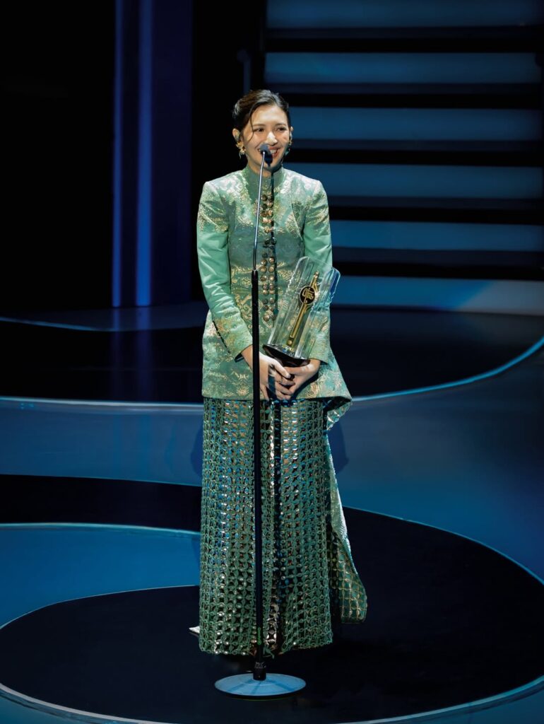 Sheila mengenakan kebaya modern berwarna hijau giok dengan aksen emas yang memantulkan cahaya panggung.