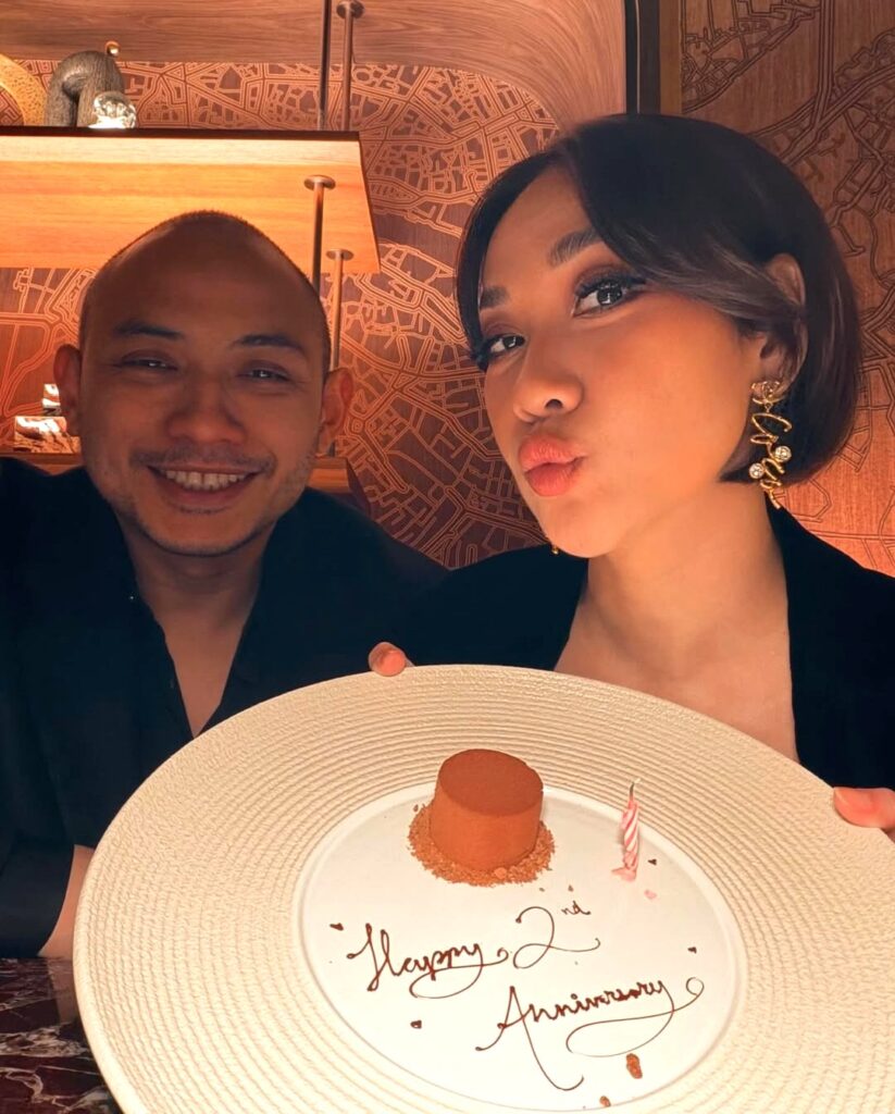 Mereka berpose bersama kue anniversary bertuliskan “Happy 2nd Anniversary” lengkap dengan lilin, menambah nuansa hangat dalam perayaan.