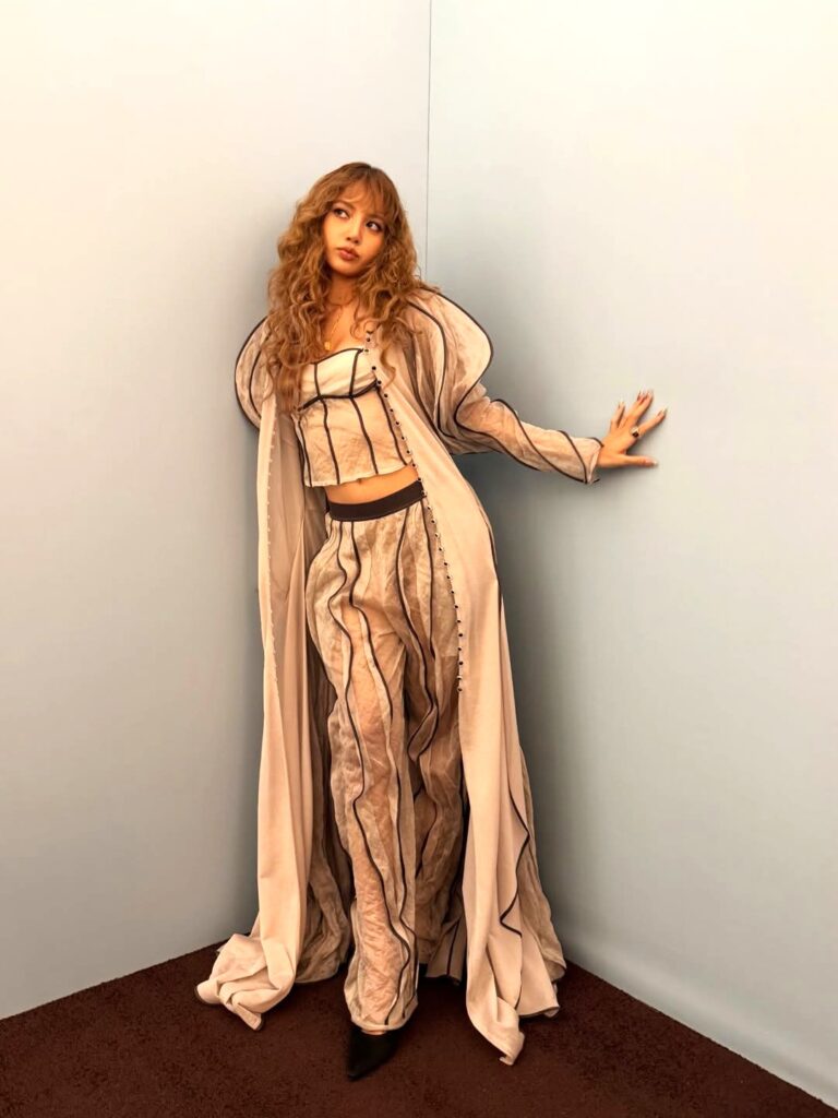 Selain rambut, busana Lisa juga tak kalah mencuri perhatian. Ia mengenakan set berbahan sheer ringan dengan aksen garis hitam kontras.